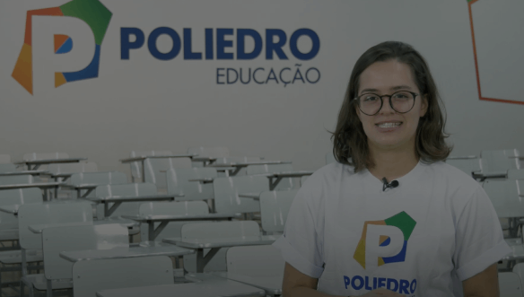 Jovem mulher sorrindo usando óculos e camiseta do Poliedro Educação, em sala de aula vazia com cadeiras e mesa ao fundo.