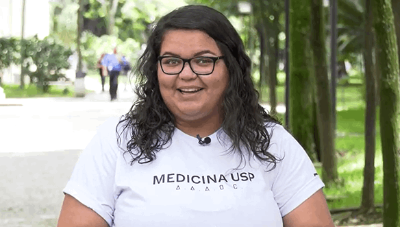Mulher sorrindo ao ar livre, usando uma camiseta branca com o texto "MEDICINA USP" e óculos, com árvores verdes ao fundo.