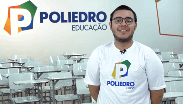 Rapaz sorrindo com barba, óculos e camiseta branca com logo do Poliedro Curso, em sala de aula com cadeiras vazias ao fundo.