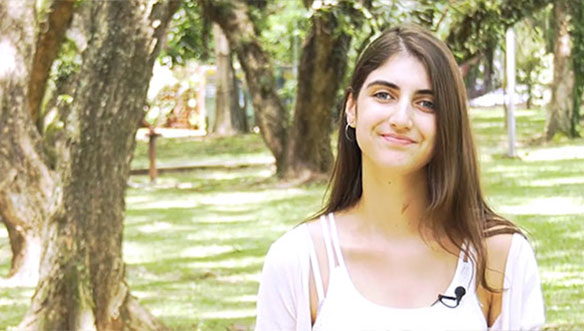 Mulher com cabelo castanho claro, usando camiseta branca, sorrindo ao ar livre com árvores ao fundo.