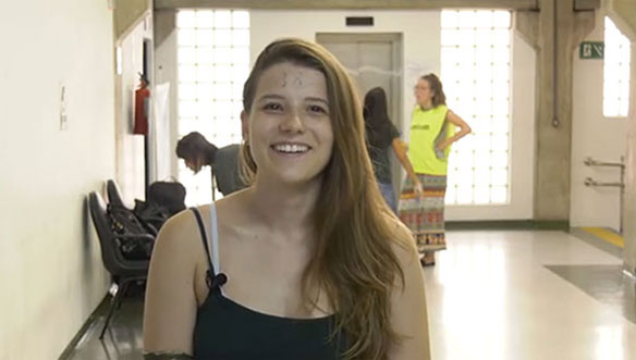 Mulher sorridente com cabelo loiro, usando roupa preta, posando em ambiente interno com bancos e pessoas ao fundo.