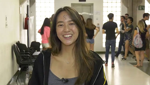 Jovem mulher sorrindo, com cabelo castanho, usando jaqueta preta e camiseta cinza, em ambiente com várias pessoas ao fundo.
