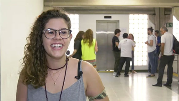 Mulher sorrindo, com óculos e cabelo cacheado, em ambiente de corredor com várias pessoas ao fundo.