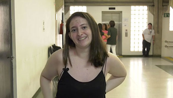 Mulher jovem com cabelo castanho, com roupa preta, sorrindo e com mãos na cintura, em ambiente interno com pessoas ao fundo.