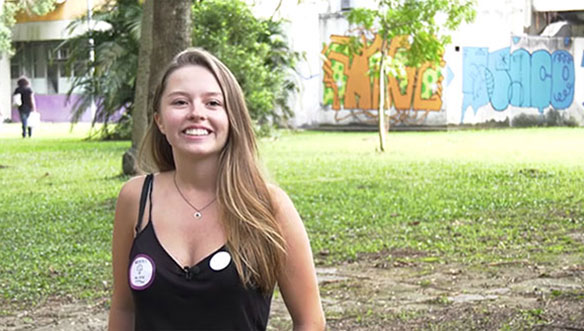 Mulher sorrindo ao ar livre, com cabelo loiro, vestindo blusa preta, em um parque com grama, árvores e grafite ao fundo.