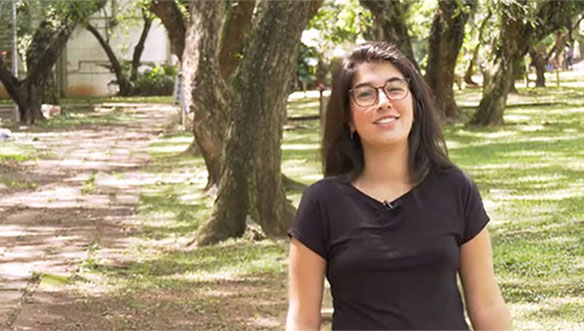 Mulher com óculos e cabelo escuro, usando camiseta preta, sorrindo ao ar livre em um parque com árvores e grama ao fundo.