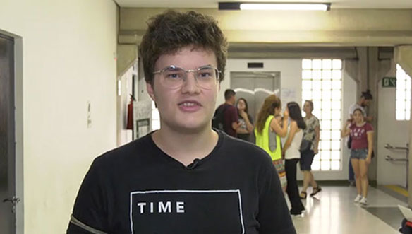 Homem jovem com óculos e cabelo castanho, usando camiseta preta, em ambiente interno com pessoas ao fundo.