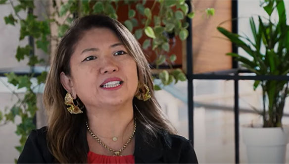 Mulher com cabelo castanho, usando brincos grandes dourados, blusa vermelha e jaqueta preta em ambiente interno.