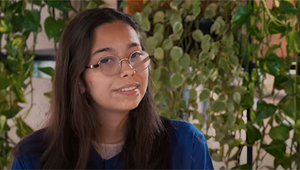 Mulher de cabelo castanho escuro, usando óculos, camiseta azul, sorrindo levemente, com plantas verdes ao fundo.