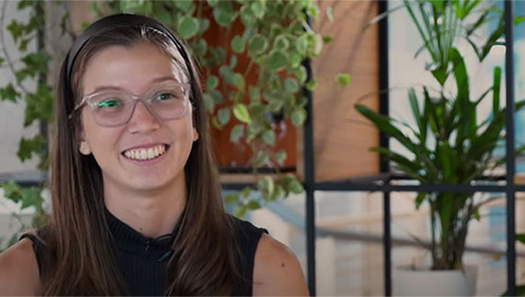 Mulher jovem sorridente com óculos e cabelo castanho, usando blusa preta, sentada em ambiente com plantas ao fundo.
