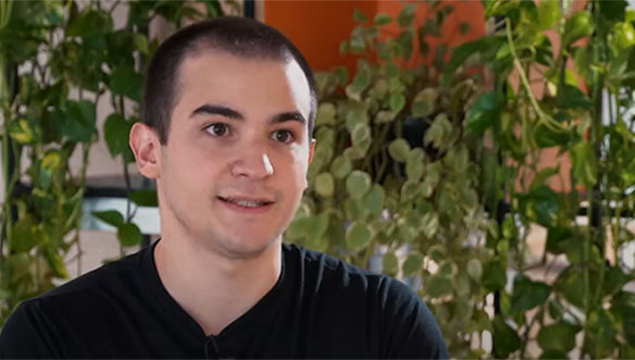 Homem com cabelo curto e escuro, usando camiseta preta, sentado em ambiente com plantas verdes ao fundo.